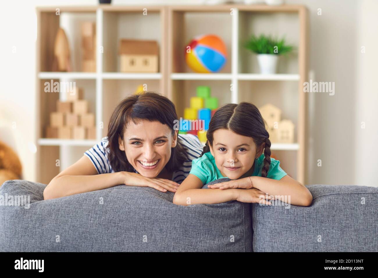 Happy mother and smiling daughter looking at camera from behind sofa at home Stock Photo