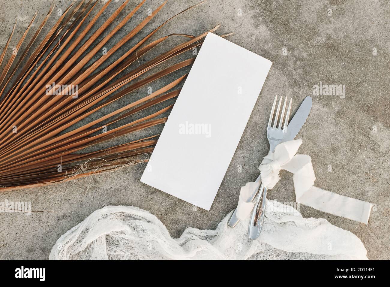 Tropical wedding ry still life. Closeup of blank menu card mock-up. Dry palm leaf, silver cutlery and muslin runner on grunge beige concrete Stock Photo