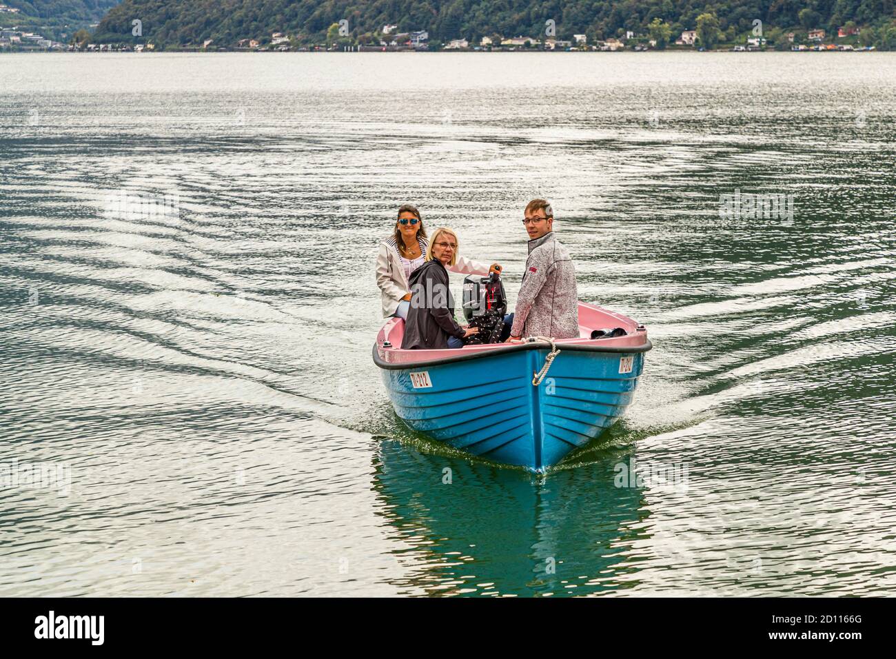 The pink-and-blue motorboat of host Gabriella Monfredini heads for the jetty with guests aboard, Lake Lugano near Melide, Ticino, Switzerland. Seen from the water, the landscape looks unfamiliar. Since 2015, the fisherman's daughter has run the culinary experience "Swiss Tavolata" from her Cantine, a former natural cellar in Melide that she converted. Stock Photo