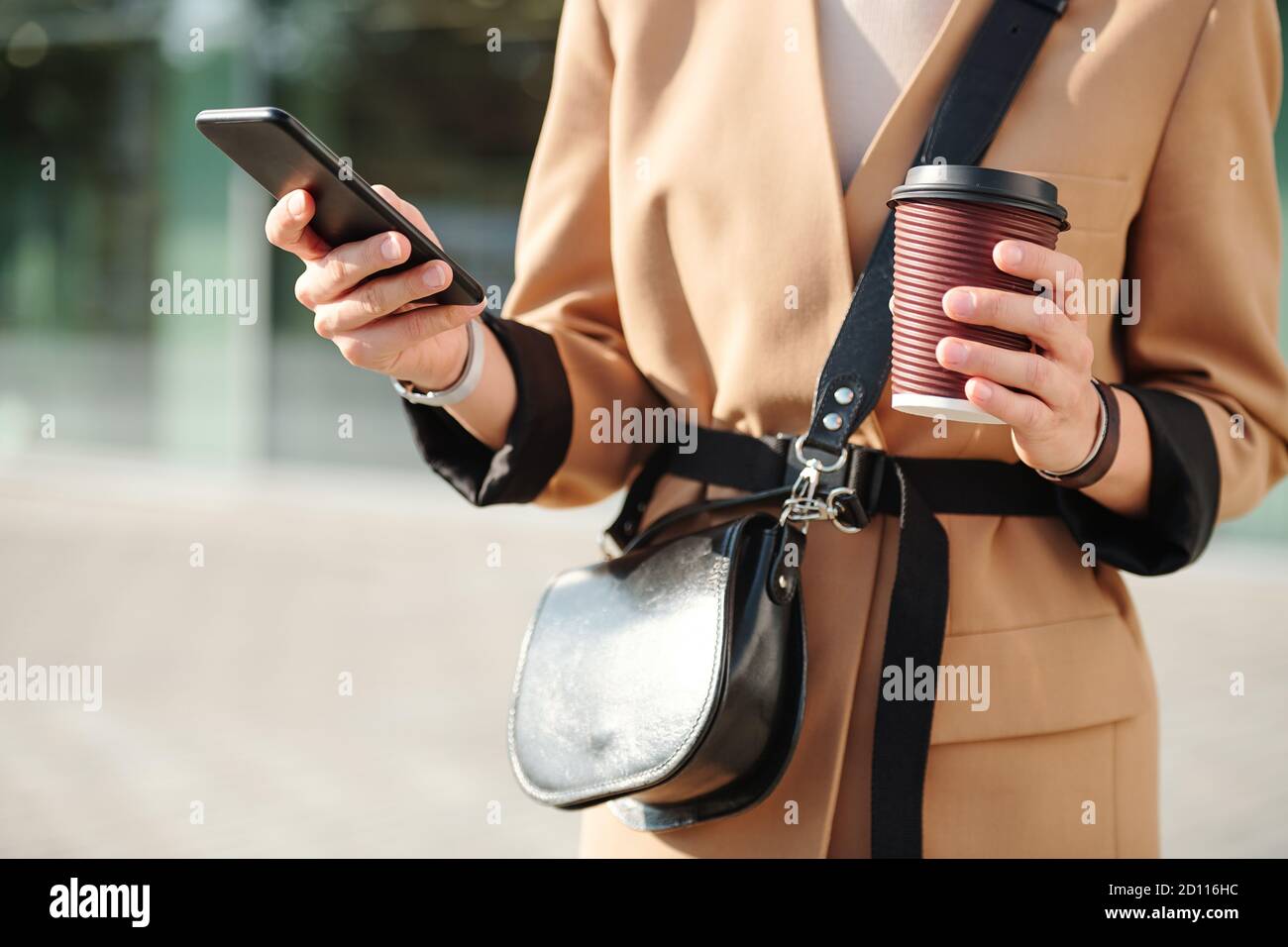 Hands of young elegant female with drink and handbag scrolling through contacts Stock Photo