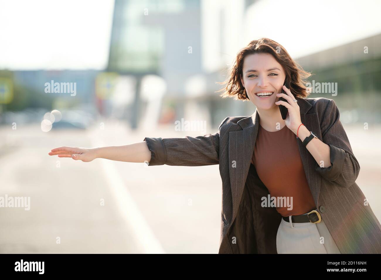 Young cheerful brunette businesswoman with smartphone by ear catching taxi Stock Photo