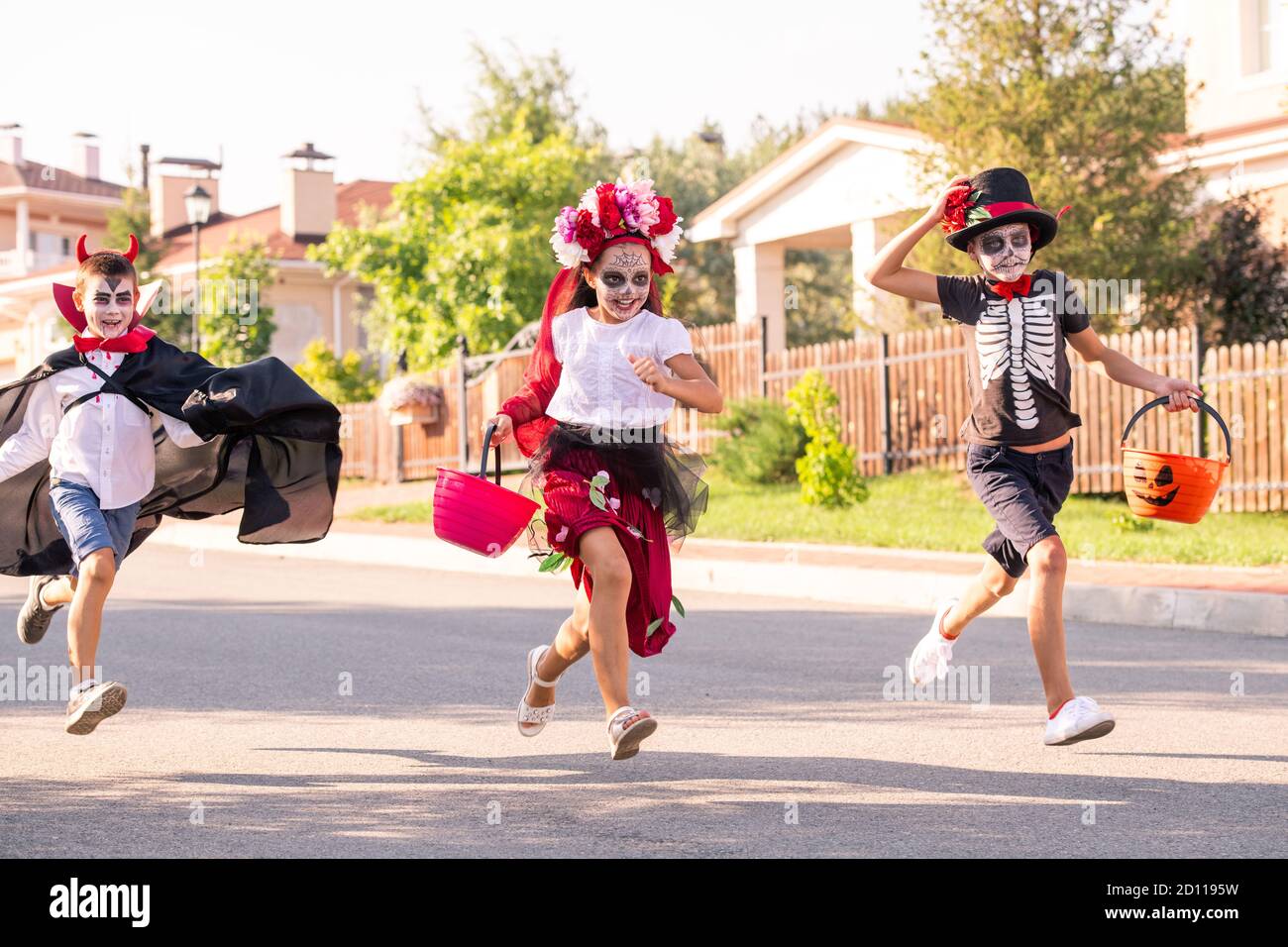 Three laughing kids in halloween costumes running down wide road on sunny day Stock Photo