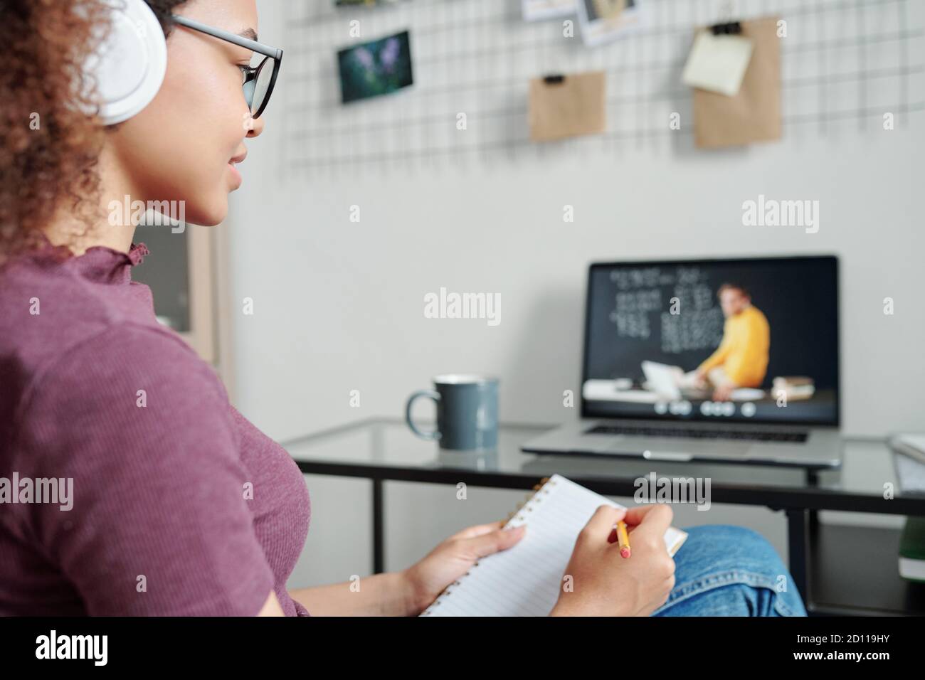 Young contemporary smiling female student making notes during online lesson Stock Photo
