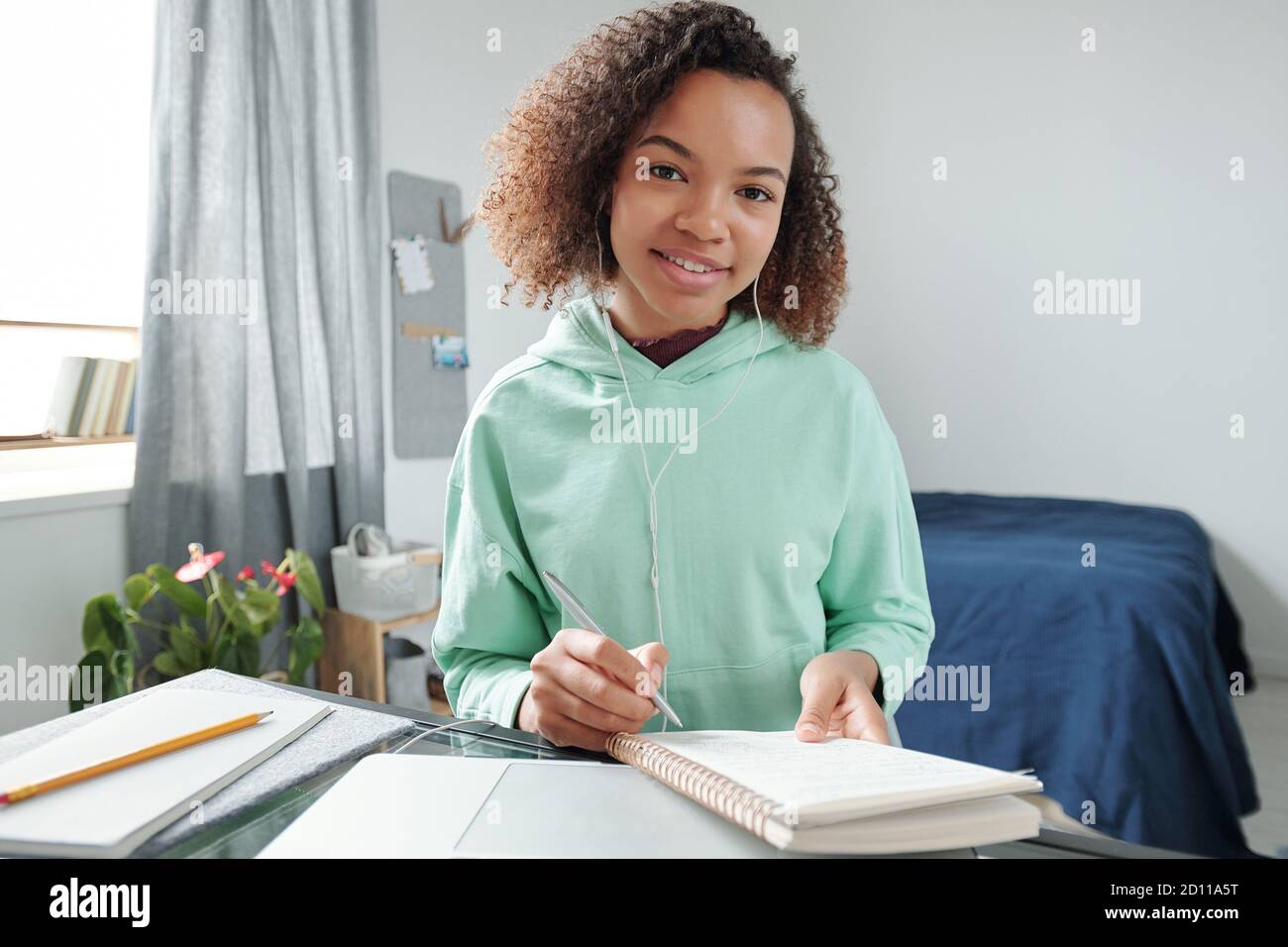 Cheerful mixed-race teenage student making notes in copybook in bedroom Stock Photo