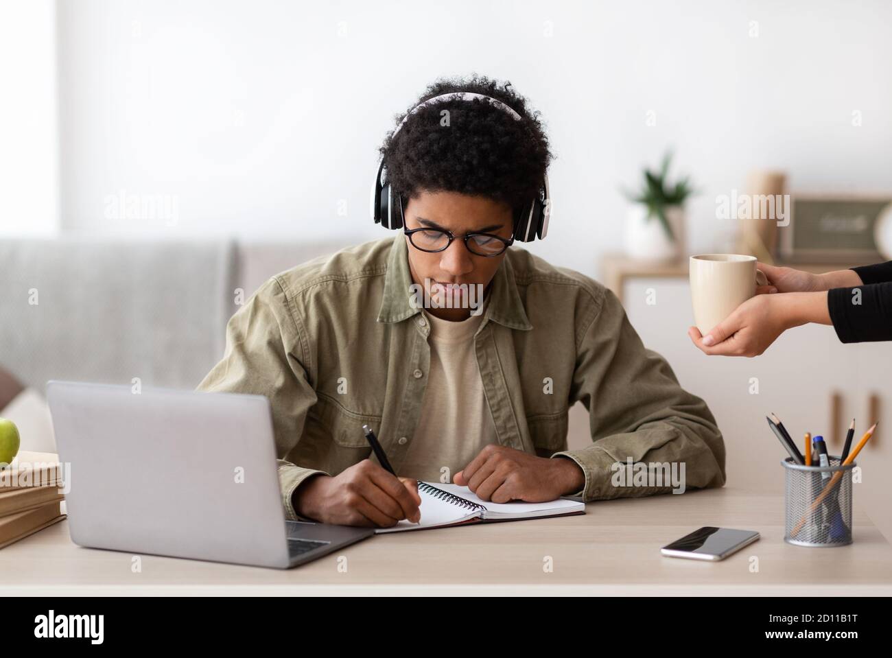Black teen guy in headphones studying online from home, making notes in copybook Stock Photo