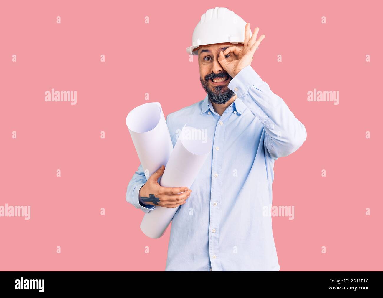 Young handsome man wearing hardhat holding paper blueprints smiling happy doing ok sign with hand on eye looking through fingers Stock Photo