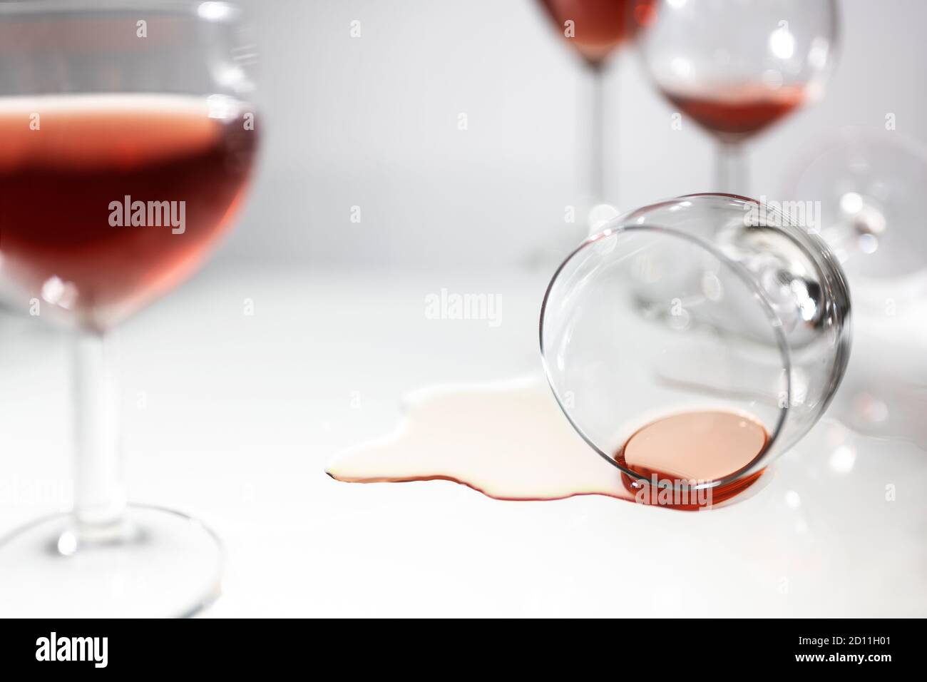 Overturned drinking glass with a puddle of red wine between blurred standing glasses, concept for party fun, alcohol addiction or burn out, light gray Stock Photo