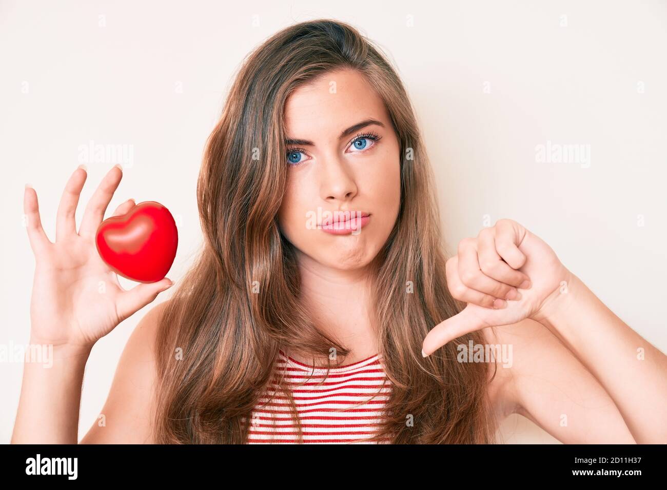 Beautiful young caucasian woman holding heart with angry face, negative sign showing dislike with thumbs down, rejection concept Stock Photo