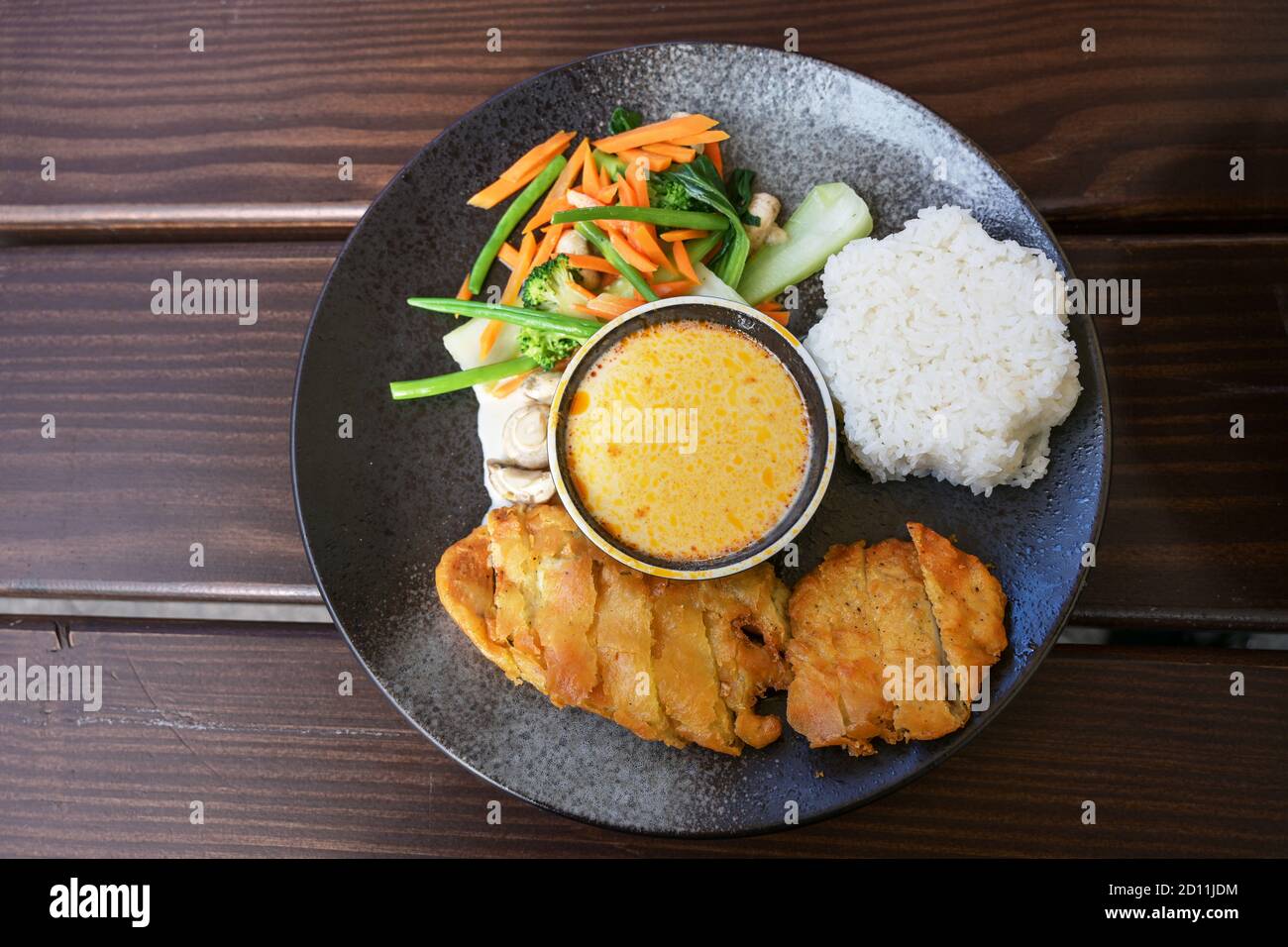 Asian dish with fried chicken, vegetables, steamed rice and coconut curry sauce on dark gray plate with chopsticks on a wooden table, high angle view Stock Photo