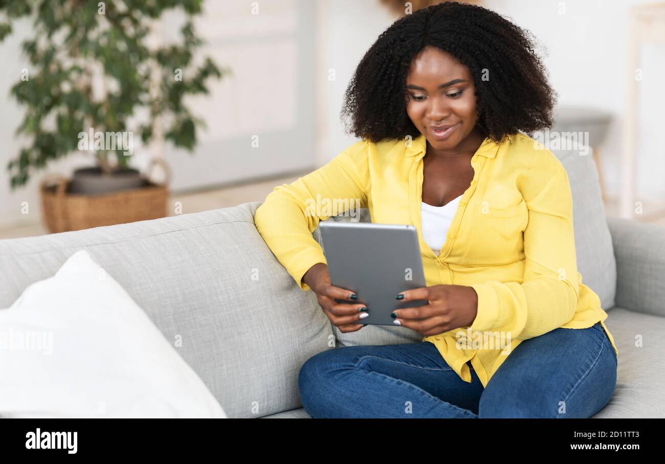 Black woman sitting on sofa, using tablet Stock Photo - Alamy