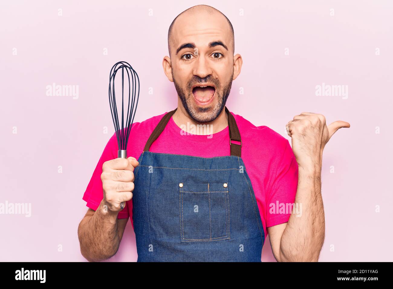 Young handsome bald man wearing apron holding whisk pointing thumb up