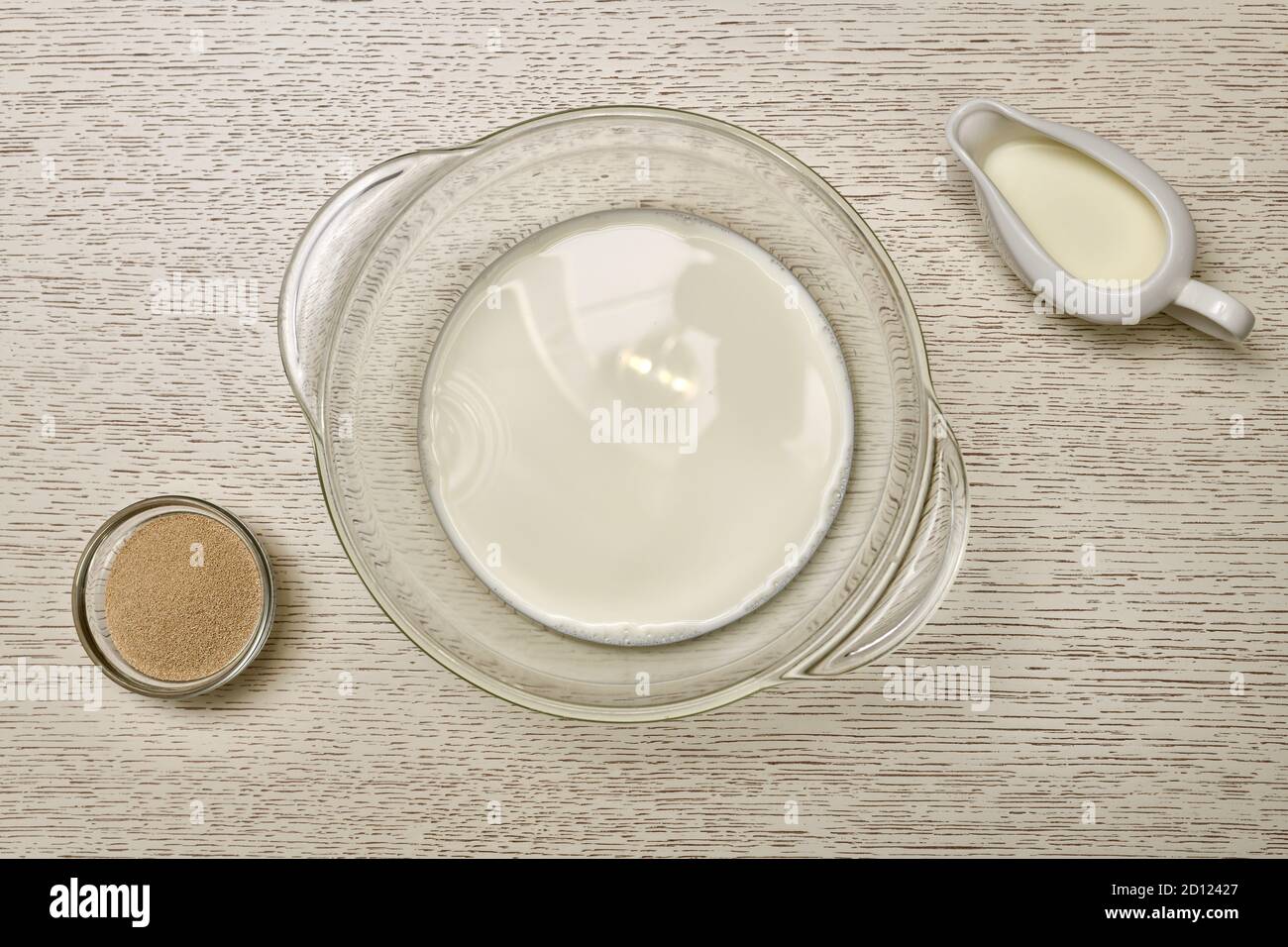 The stage of preparation yeast dough. Transparent pan with milk. Next to the yeast and creamer. On a cream countertop. Stock Photo