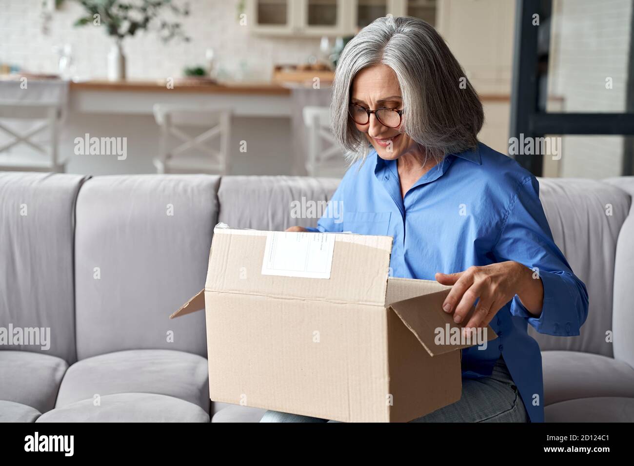 Smiling older woman unpacking parcel postal delivery box at home on couch. Stock Photo