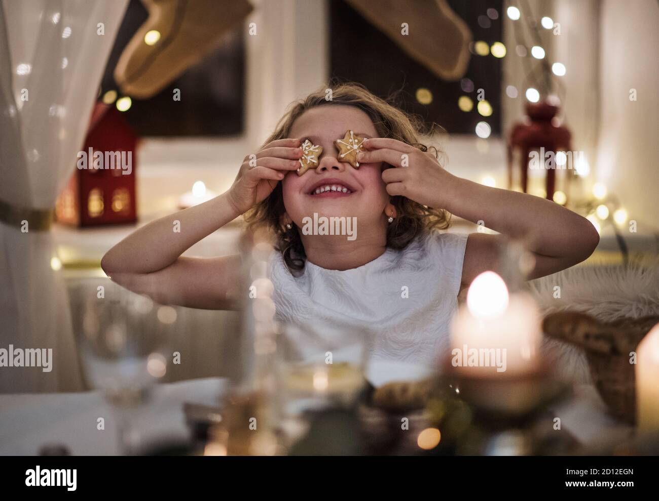 Small girl sitting indoors at Christmas, covering eyes with gingerbread biscuit. Stock Photo