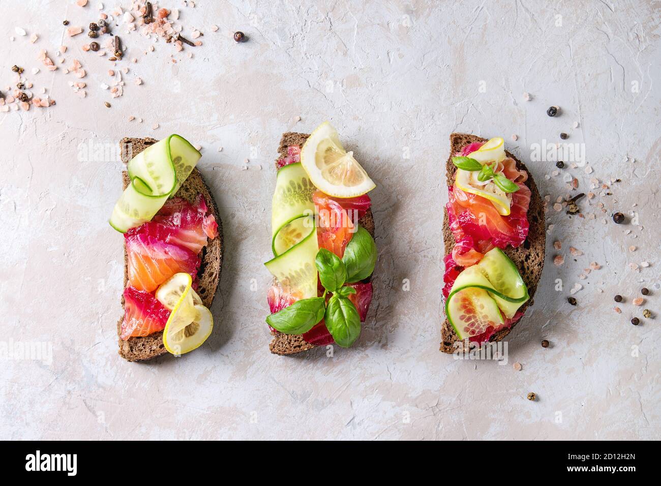 Sliced beetroot marinated salmon sandwiches with rye bread, cucumber, basil and lemon over grey texture background. Top view, space Stock Photo