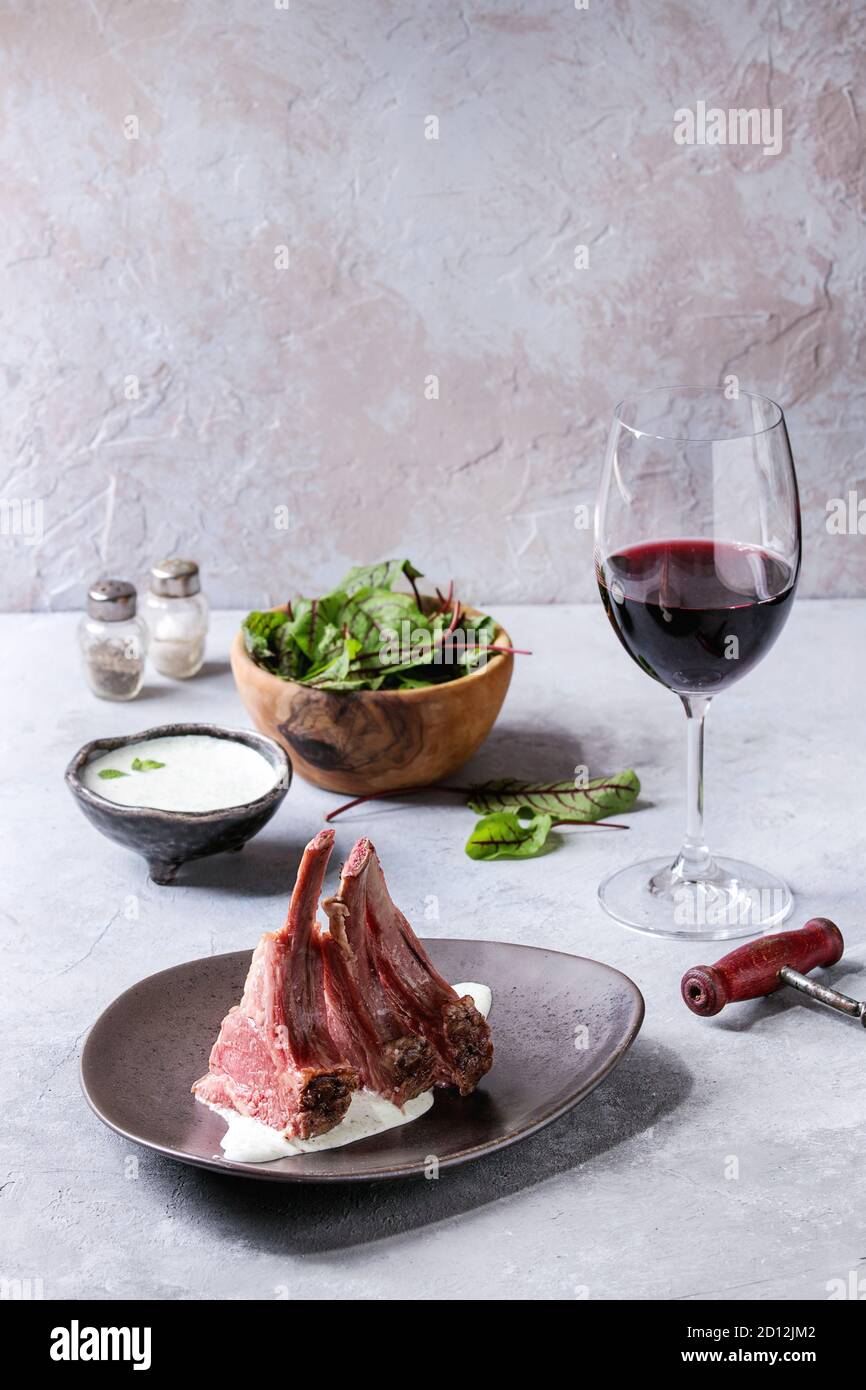 Grilled sliced rack of lamb with yogurt mint sauce served in ceramic plate with green salad young beetroot leaves, glass of red wine over grey texture Stock Photo