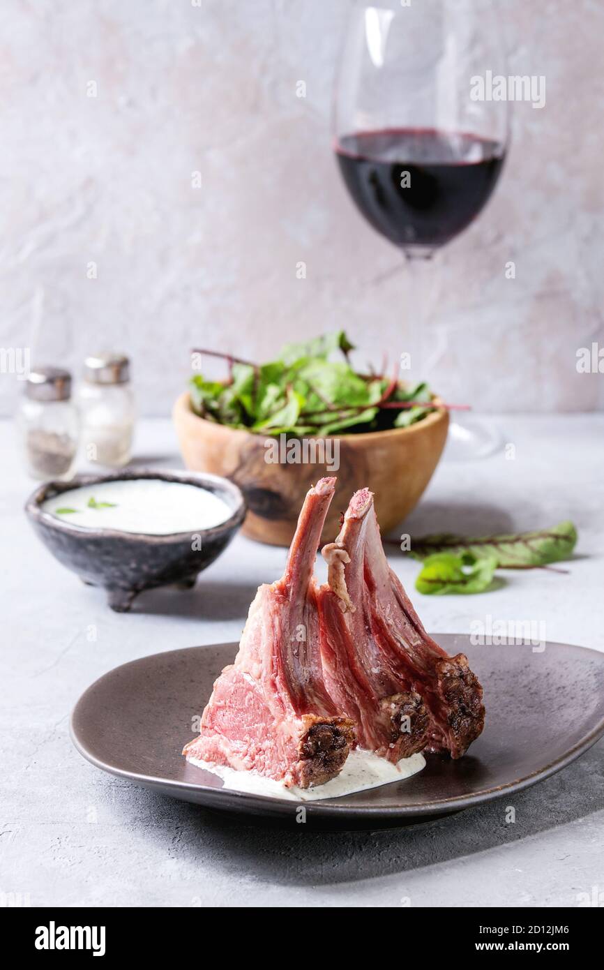 Grilled sliced rack of lamb with yogurt mint sauce served in ceramic plate with green salad young beetroot leaves, glass of red wine over grey texture Stock Photo