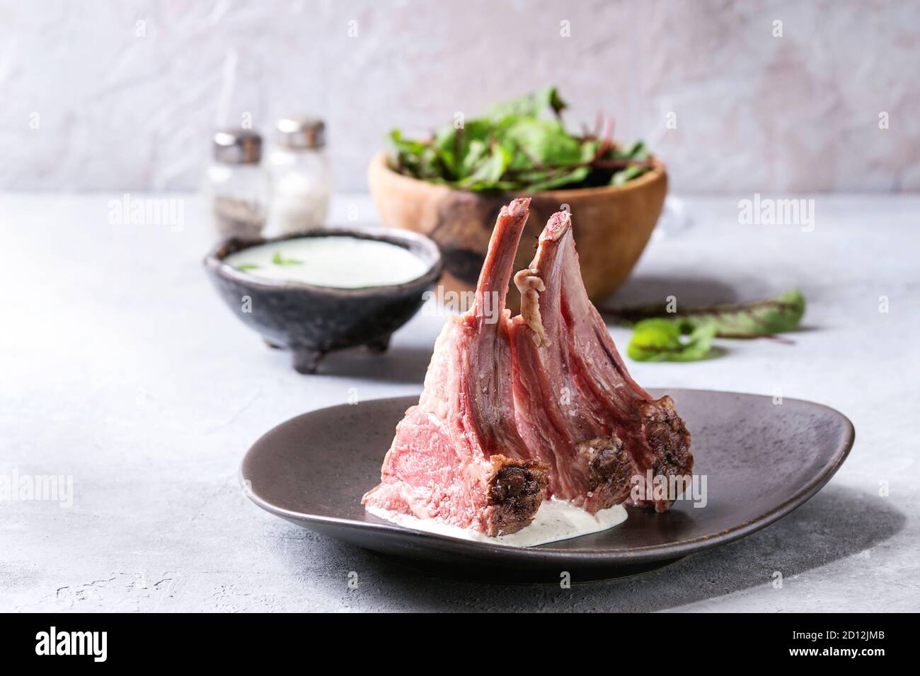 Grilled sliced rack of lamb with yogurt mint sauce served in ceramic plate with green salad young beetroot leaves over grey texture table. Stock Photo