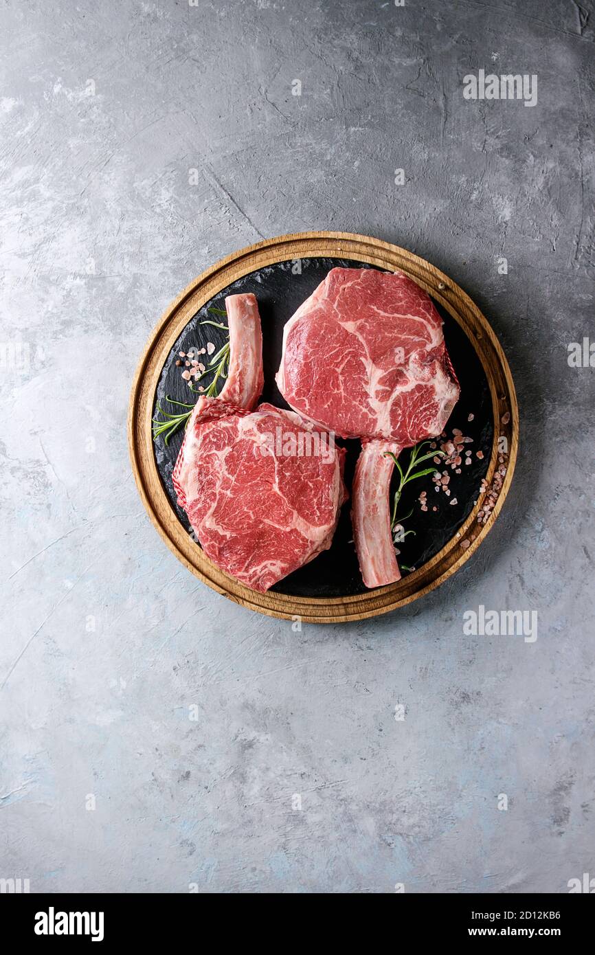 Raw uncooked black angus beef tomahawk steaks on bones served with salt and pepper on round wooden slate cutting board over grey texture background. T Stock Photo