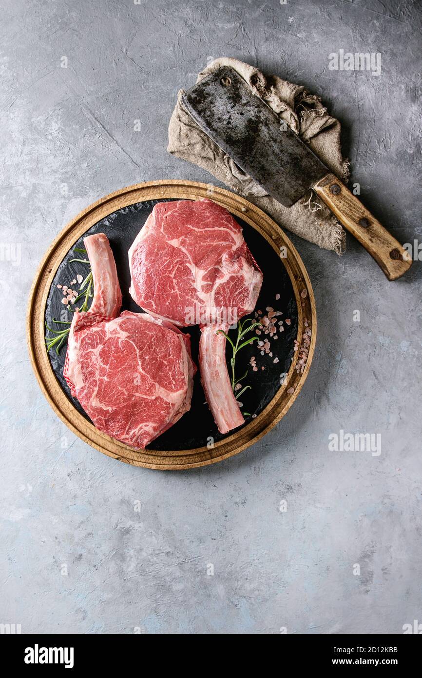 Raw uncooked black angus beef tomahawk steaks on bones served with salt, pepper, vintage butcher cleaver on round wooden slate cutting board over grey Stock Photo