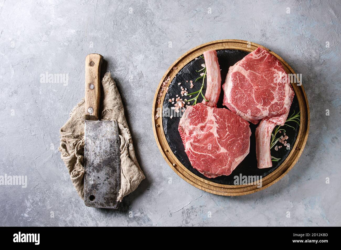 Raw uncooked black angus beef tomahawk steaks on bones served with salt, pepper, vintage butcher cleaver on round wooden slate cutting board over grey Stock Photo