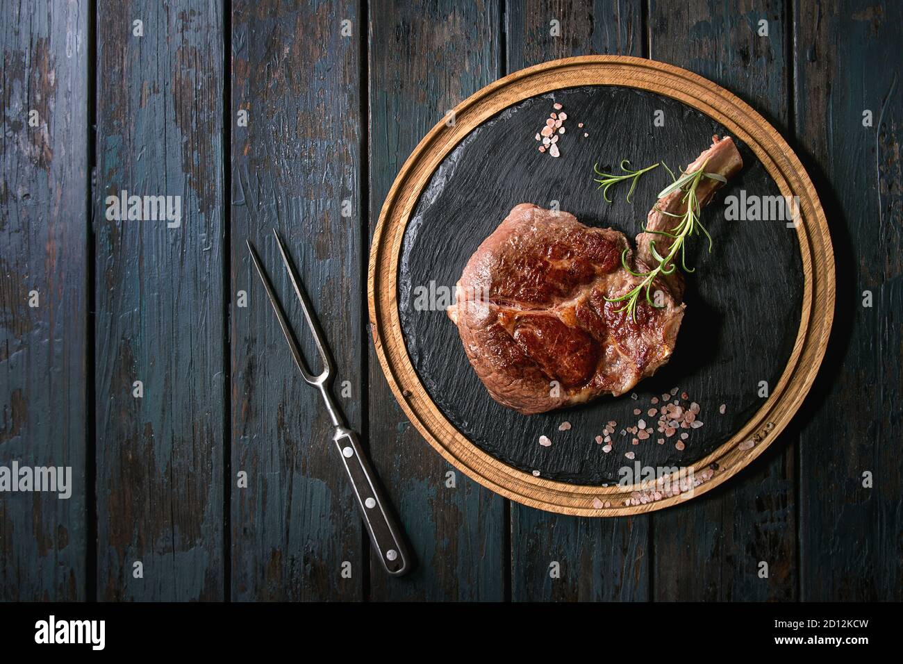 Grilled black angus beef tomahawk steak on bone served with salt, pepper and rosemary on round wooden slate cutting board with meat fork over dark woo Stock Photo