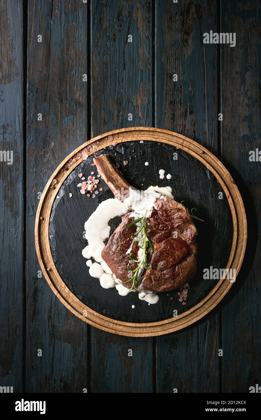 Grilled black angus beef tomahawk steak on bone served with salt, pepper, rosemary and white sauce on round wooden slate cutting board over dark woode Stock Photo