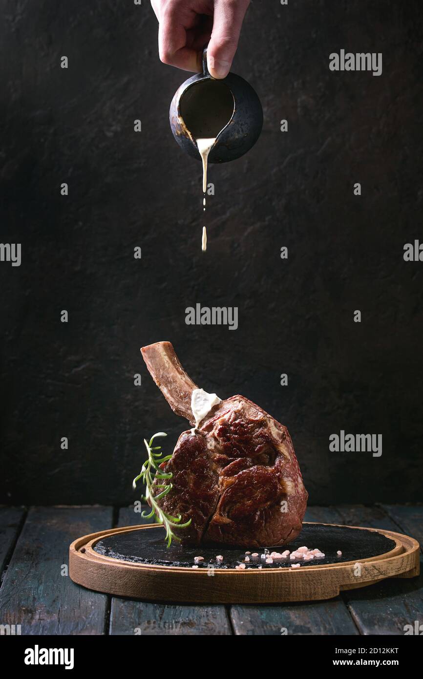 Grilled black angus beef tomahawk steak on bone served with salt, pepper, rosemary, flowing white sauce on round slate cutting board over dark wooden Stock Photo