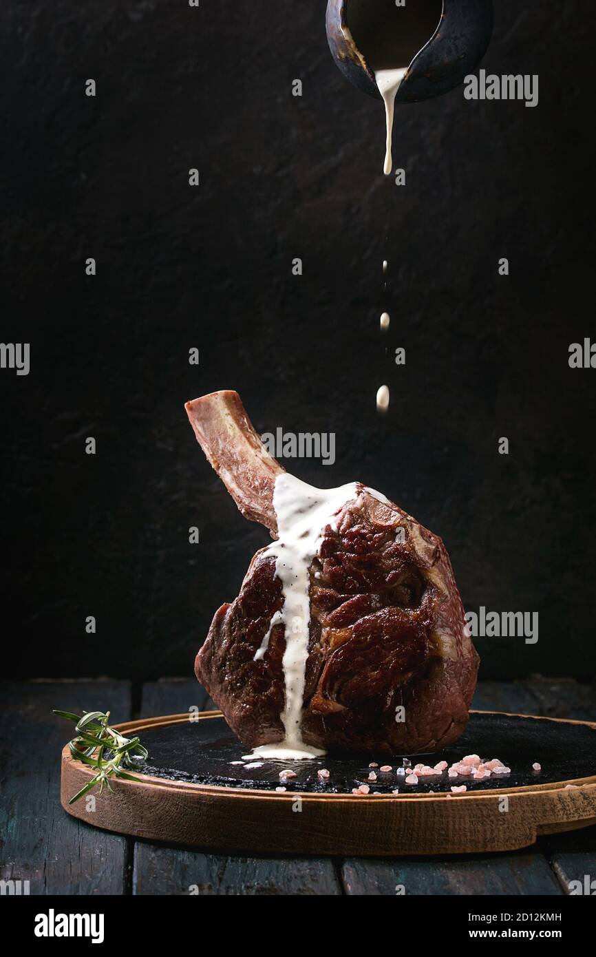 Grilled black angus beef tomahawk steak on bone served with salt, pepper, rosemary, flowing white sauce on round slate cutting board over dark wooden Stock Photo