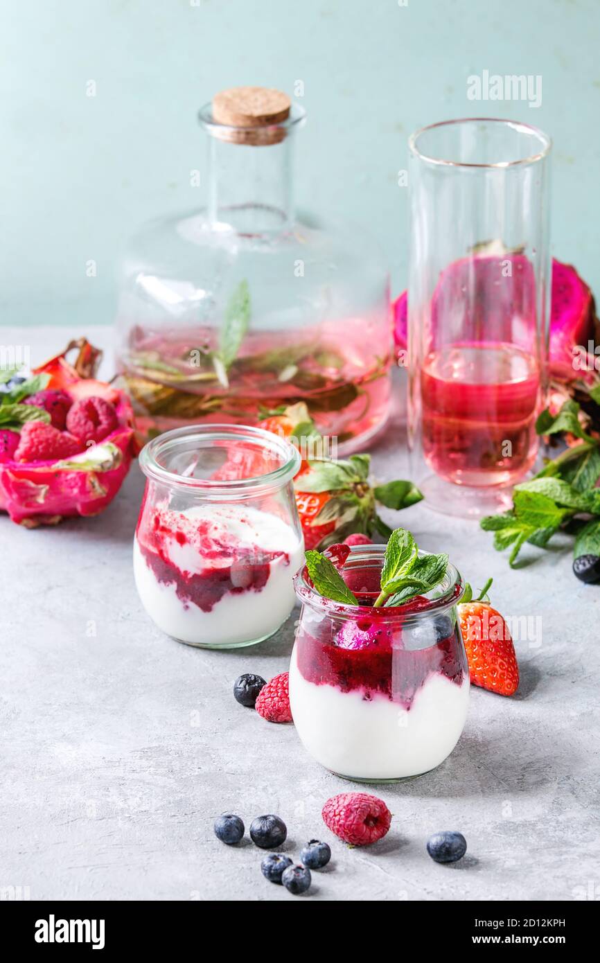 Jars of natural white yogurt with berry sauce, fruit salad with pink dragon fruit, berries and mint, served with bottle of lemonade on grey table. Hea Stock Photo