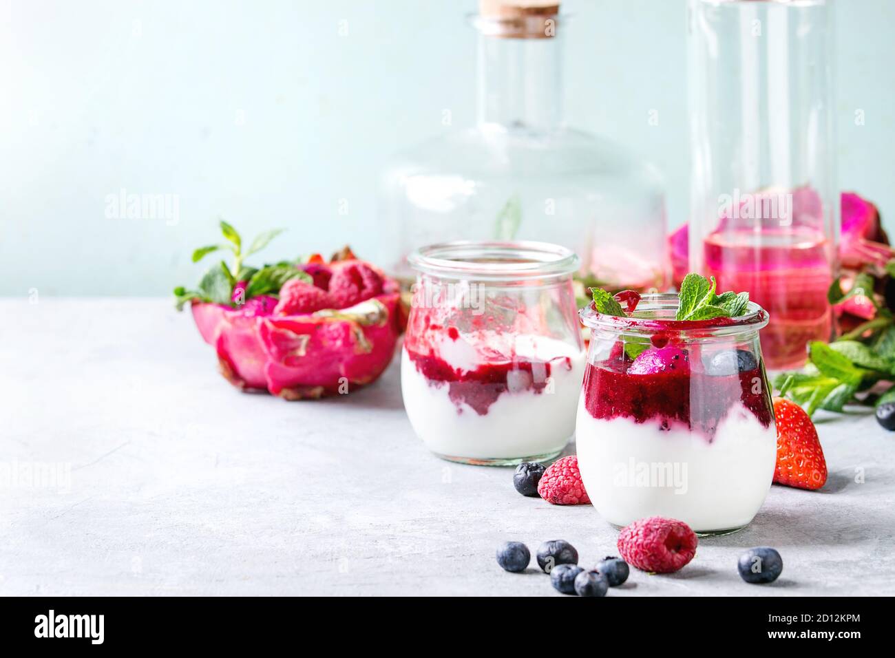 Jars of natural white yogurt with berry sauce, fruit salad with pink dragon fruit, berries and mint, served with bottle of lemonade on grey table. Hea Stock Photo