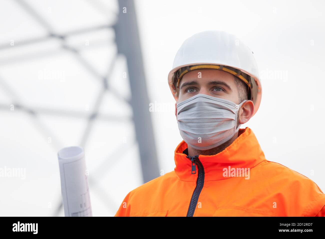 Young engineer or architect with face mask and plan in his hand wearing protective work wear and hard hat - selective focus Stock Photo