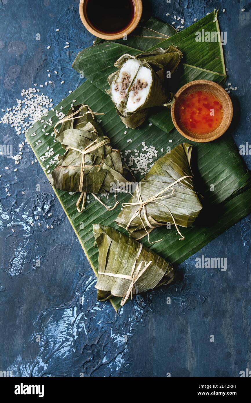 Asian rice piramidal steamed dumplings from rice tapioca flour with meat filling in banana leaves. Ingredients and sauces above over blue texture back Stock Photo