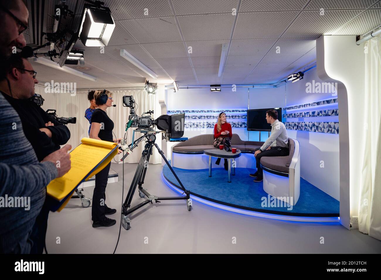 A TV show being filmed in a studio. The presenters are sitting on the studio sofa and talking to each other while the camera crew films them with film Stock Photo