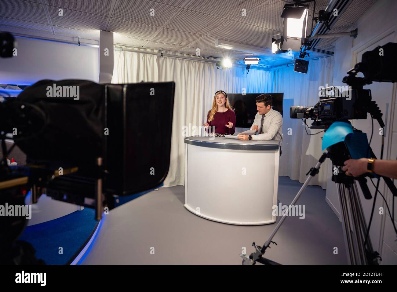 A TV show in the process of being filmed in a studio. The presenters are sitting at the studio desk, talking to the camera. Stock Photo