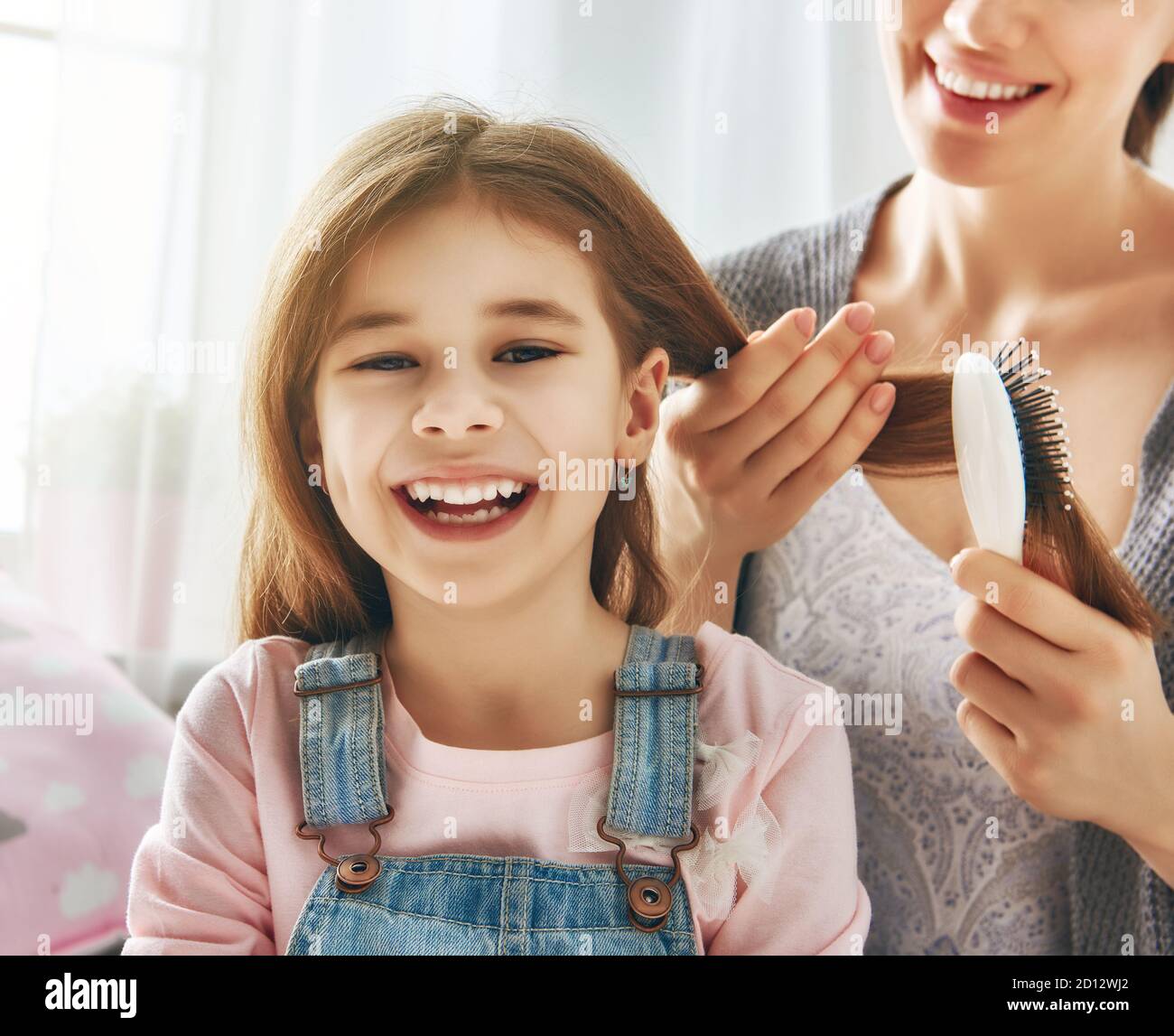 Happy loving family. Mother is combing her daughter's hair sitting on the bed in the room. Stock Photo