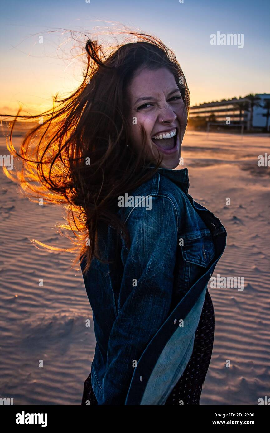 Young woman portrait on a beach sunset walk in autumn. Lateral shot. Rocking vibes at camera, hair in the air. Stock Photo