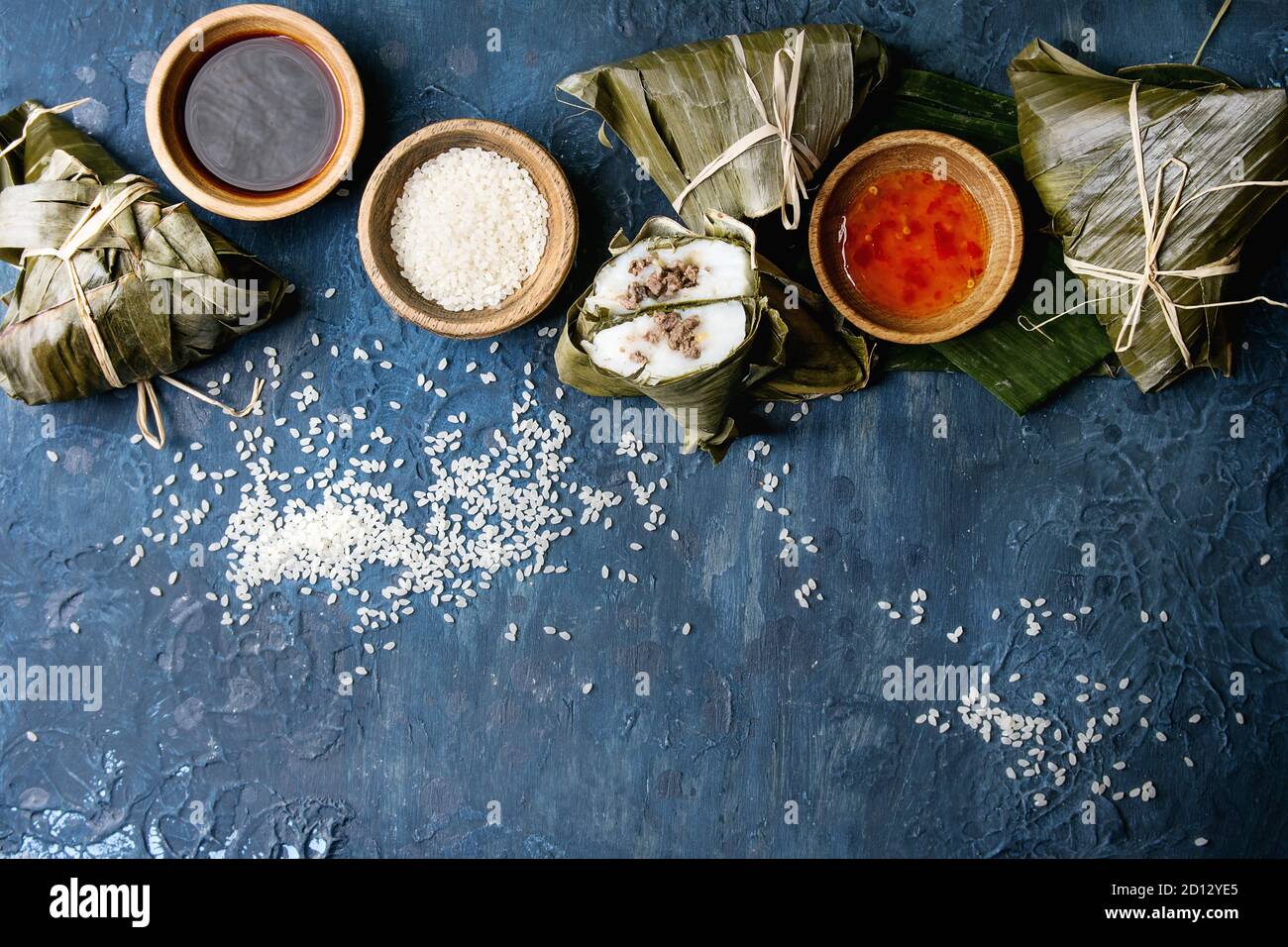 Asian rice piramidal steamed dumplings from rice tapioca flour with meat filling in banana leaves. Ingredients and sauces above over blue texture back Stock Photo