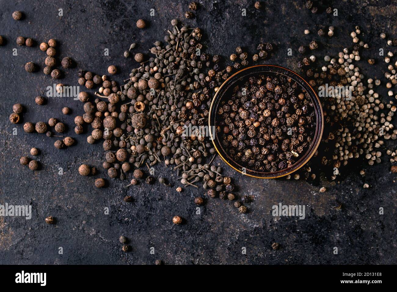 Variety of different black peppers allspice, pimento, monks pepper, peppercorns and ground powder in tin can over old black iron texture background. T Stock Photo