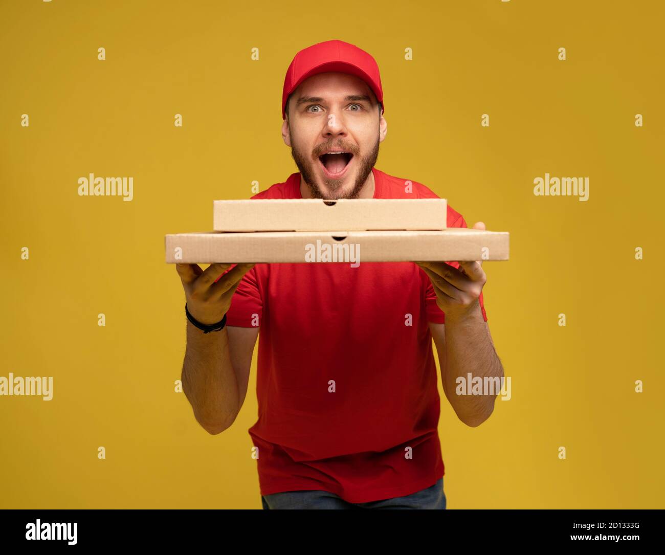Happy man from delivery service in red t-shirt and cap giving food order and holding pizza box isolated over yellow Stock Photo