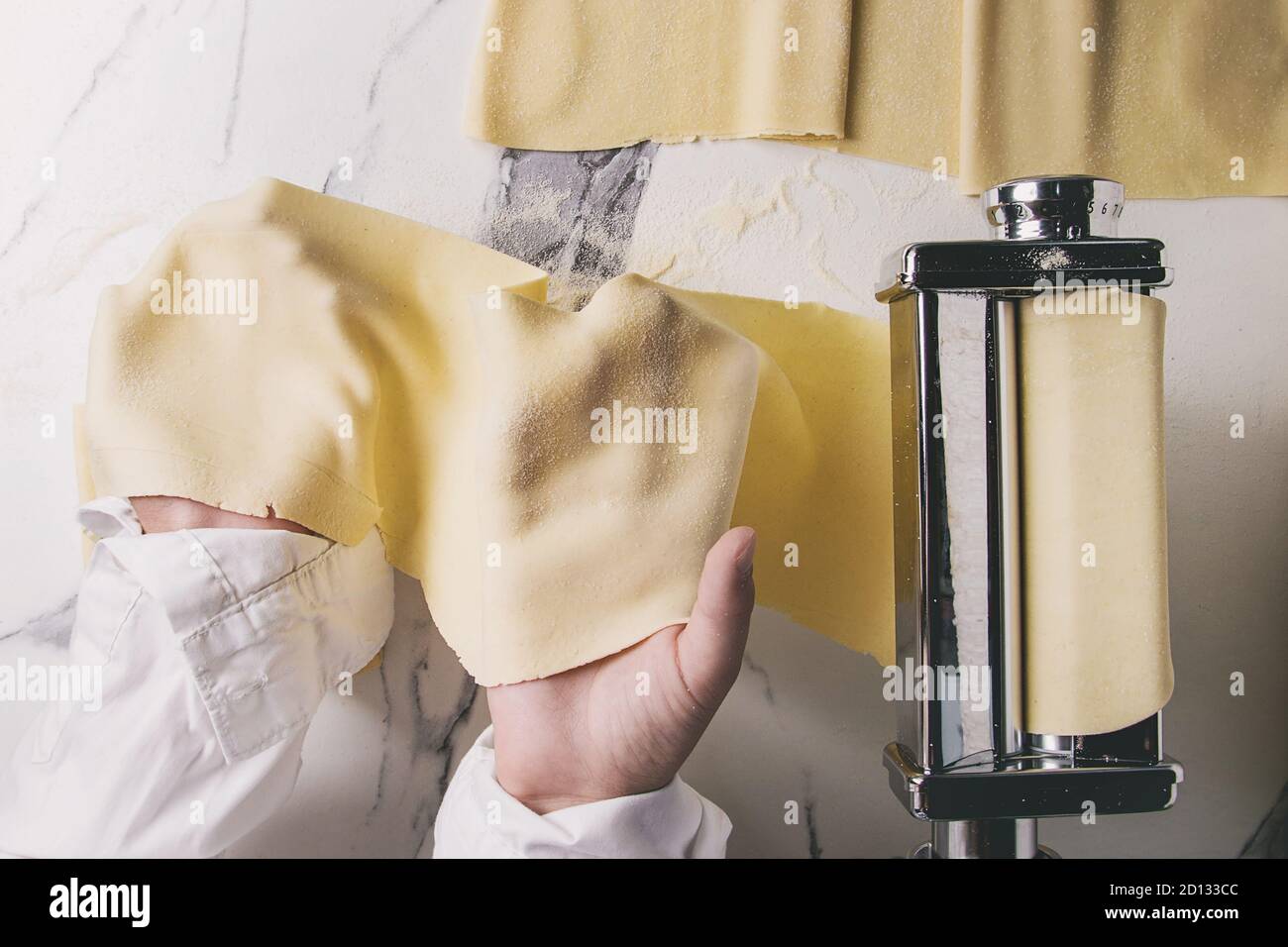 Rolled dough for homemade italian uncooked pasta lasagna from pasta machine maker in hands with semolina flour over white marble texture background. F Stock Photo