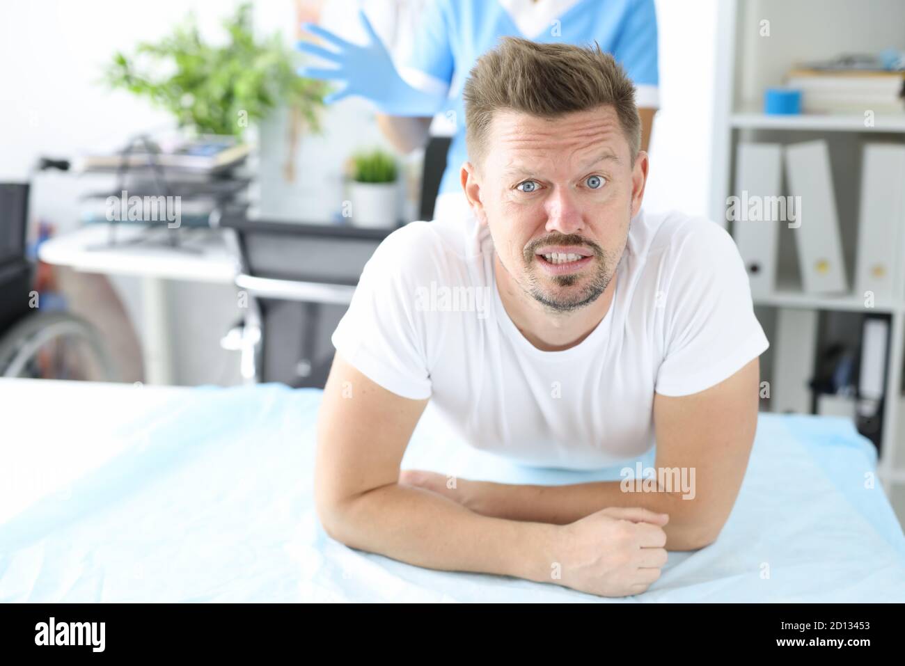 Young male patient lies on couch on stomach with doctor wearing rubber glove in background Stock Photo
