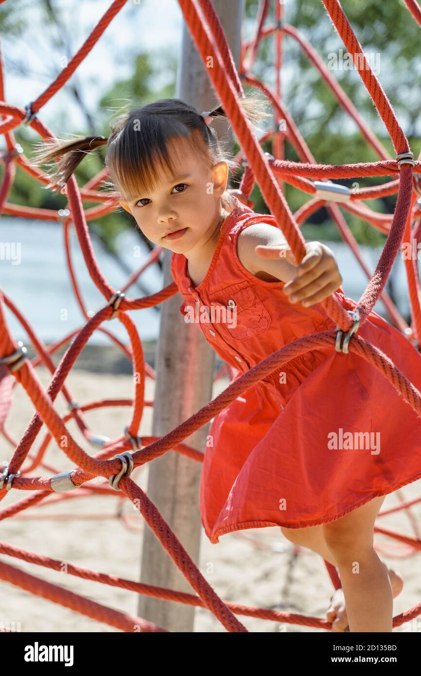 little girl in a red dress perch on a web rope-ladder structure in a children's playground for fun climbing. Stock Photo