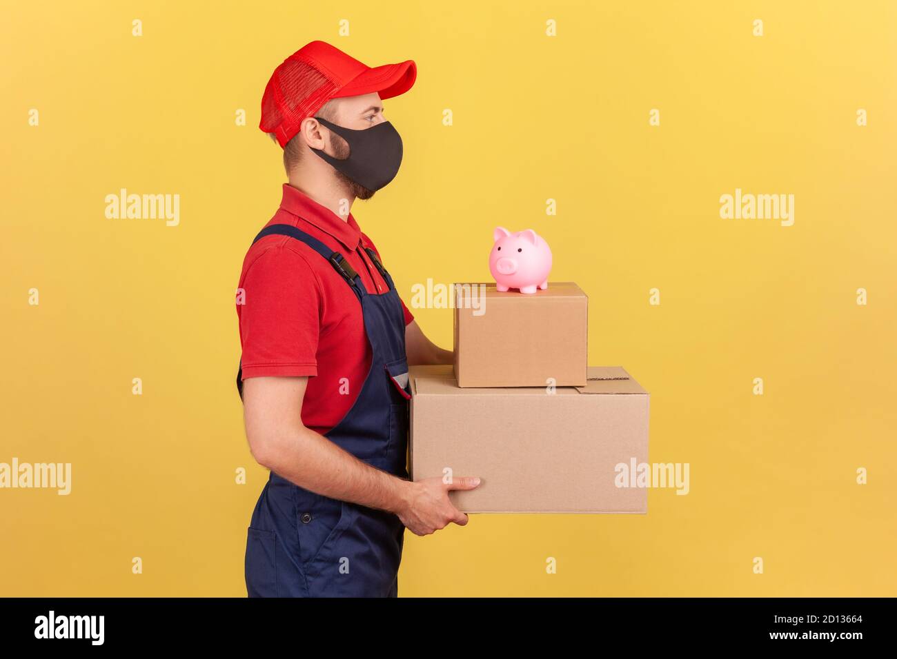 Profile portrait courier in uniform and protective mask holding cardboard parcels with piggy bank on top, save money ordering delivery, advertising. I Stock Photo