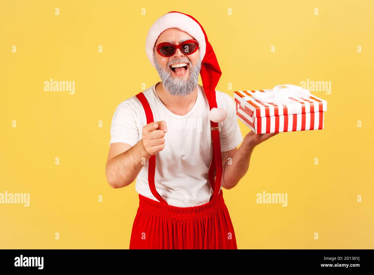 Positive excited elderly man in santa claus hat and stylish sunglasses pointing finger at you holding gift box, preparing holiday presents. Indoor stu Stock Photo