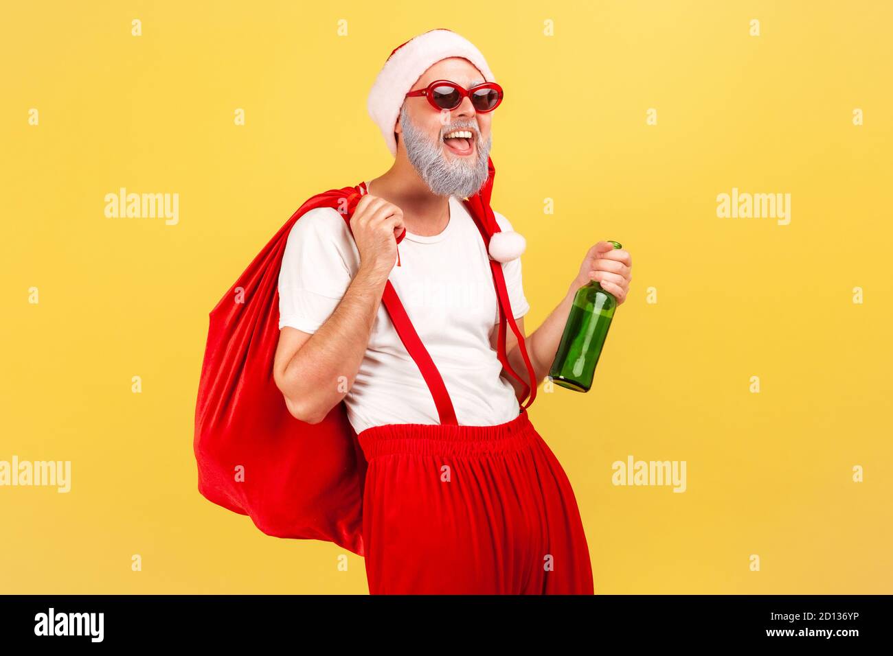 Happy elderly man in santa claus hat, trendy sunglasses and pants with suspenders holding bottle with alcohol and big red bag, celebration, bad habit. Stock Photo