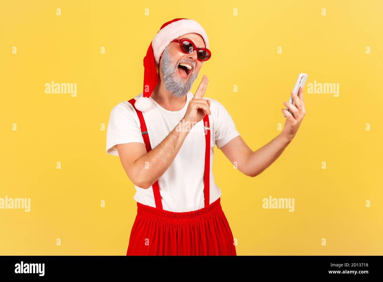Smiling positive elderly man in santa claus hat showing peace gesture talking via video call on his smartphone, blogger communicating with subscribers Stock Photo