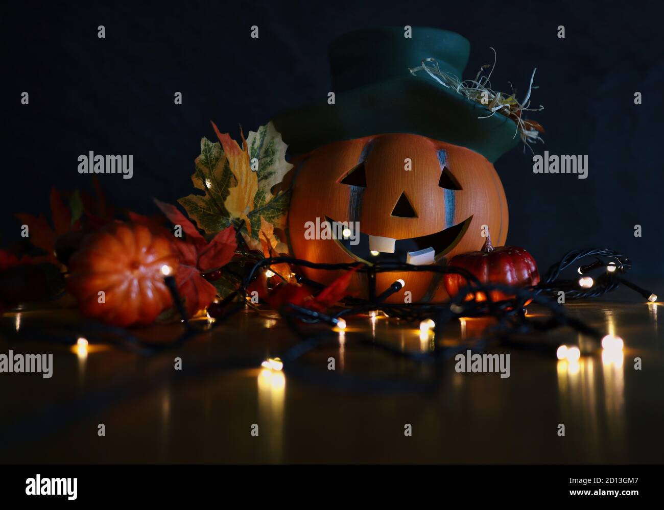 Ceramic Halloween Orange Pumpkin with Green Hat and Smile with Artificial Autumn Leaves and Blinking Lights. Moody Autumn and Halloween Vibes. Stock Photo
