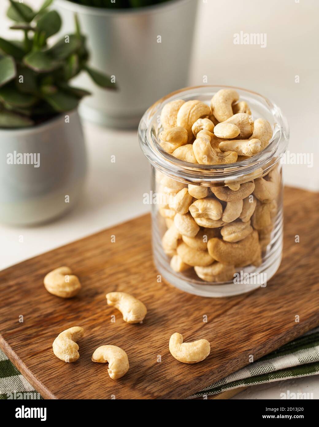 A lot of cashew nuts in a glass container on a wooden Board on a light background. green plants in pots. Stock Photo
