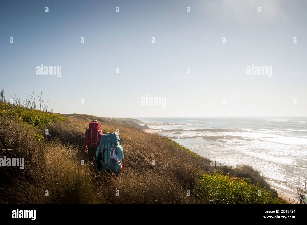 Two hikers walk the trail along the dunes and cliffs with tall vegetation, standing aside a beach landscape on late evening Stock Photo